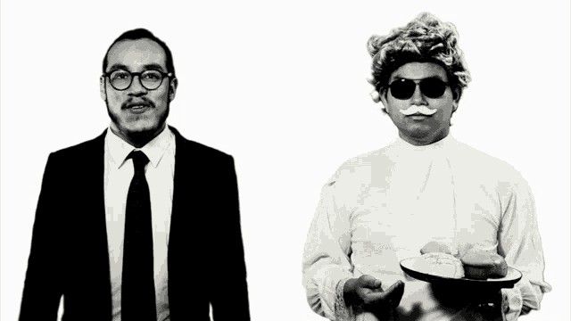 a man in a suit and tie stands next to a man in a wig and sunglasses holding a tray of cupcakes