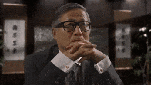 a man wearing glasses and a suit has his hands folded in front of a wall with chinese writing on it