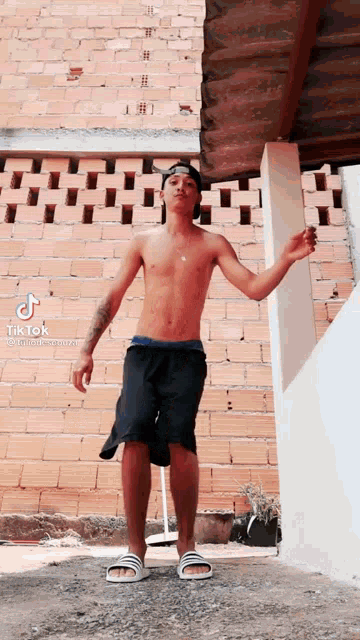 a shirtless man is standing in front of a brick wall with his arms outstretched .