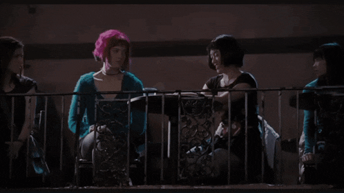 a woman with pink hair stands in front of a man 's face with an exit sign in the background