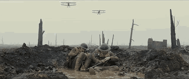 two soldiers are laying in a muddy trench with planes flying in the background
