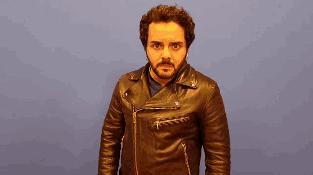 a man with a beard is wearing a leather jacket and making a surprised face