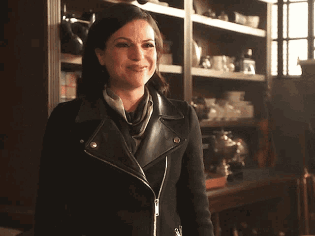 a woman wearing a black leather jacket and scarf smiles in front of a bookshelf