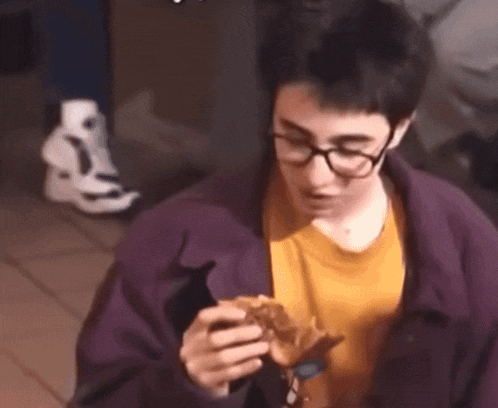 a man wearing glasses and a purple jacket is eating a sandwich .