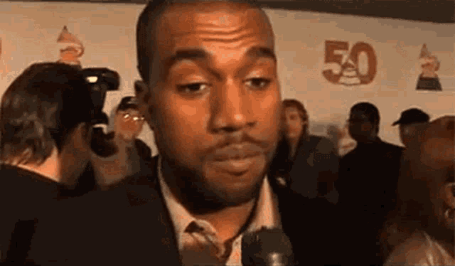 a man in a suit and tie is talking into a microphone while standing in front of a crowd .