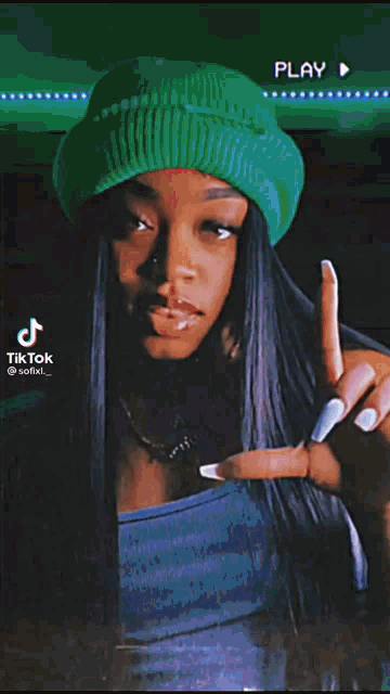 a woman wearing a green beanie and a blue top is making a peace sign .