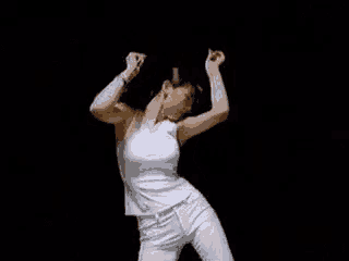 a woman in a white tank top and white pants is dancing in a dark room .