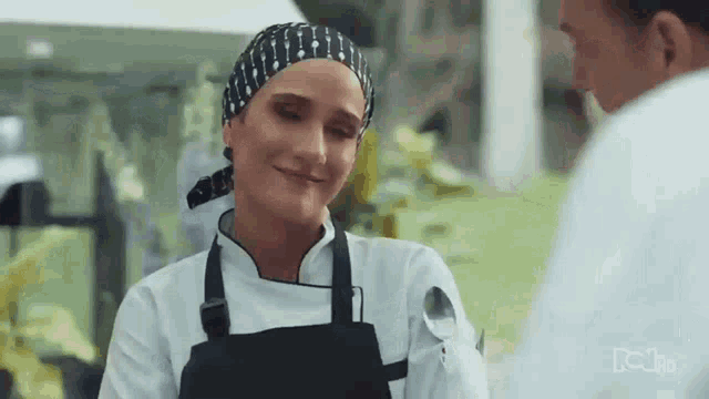 a woman in a chef 's uniform is smiling and talking to a man in a kitchen .