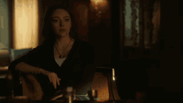 a woman sits at a table in a dark room with a necklace around her neck
