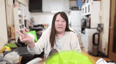 a woman with long hair is sitting on a couch in a messy room giving the peace sign .