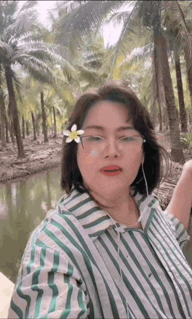 a woman wearing glasses and a flower in her hair is taking a selfie in front of palm trees .