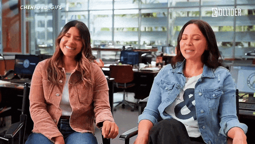 two women are sitting next to each other and laughing in an office .