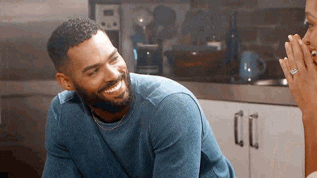 a man with a beard wearing a blue sweater is smiling in a kitchen