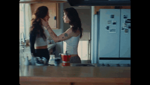 a woman with a tattoo on her arm is touching another woman 's face in the kitchen