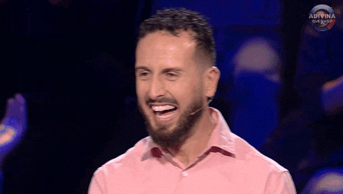 a man with a beard wearing a pink shirt is laughing on a tv show
