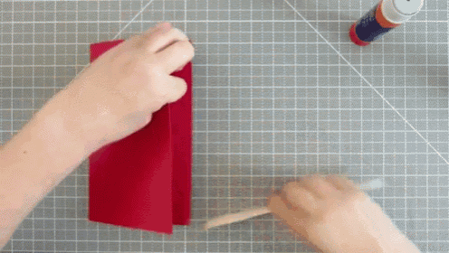 a person is holding a piece of red paper on a cutting board next to a stick of glue .