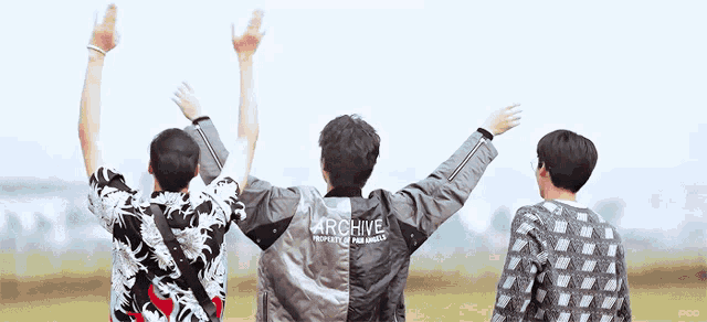 three men are standing in a field with their arms in the air and one of them has a jacket that says archive on it