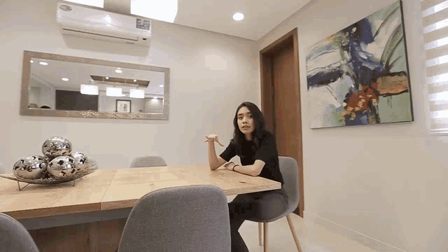 a woman sits at a table in a dining room with a painting on the wall