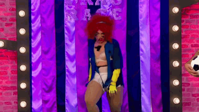 a drag queen is standing in front of a purple curtain with a soccer ball in her hand .
