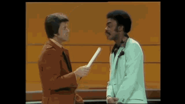 two men in suits are standing next to each other and talking . one of the men is holding a piece of paper .