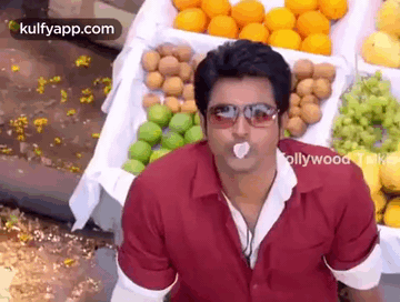 a man wearing sunglasses is sitting in front of a fruit stand with a piece of gum in his mouth .