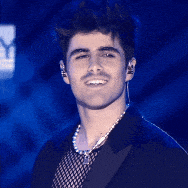 a man wearing a necklace and ear buds smiles in front of a blue background