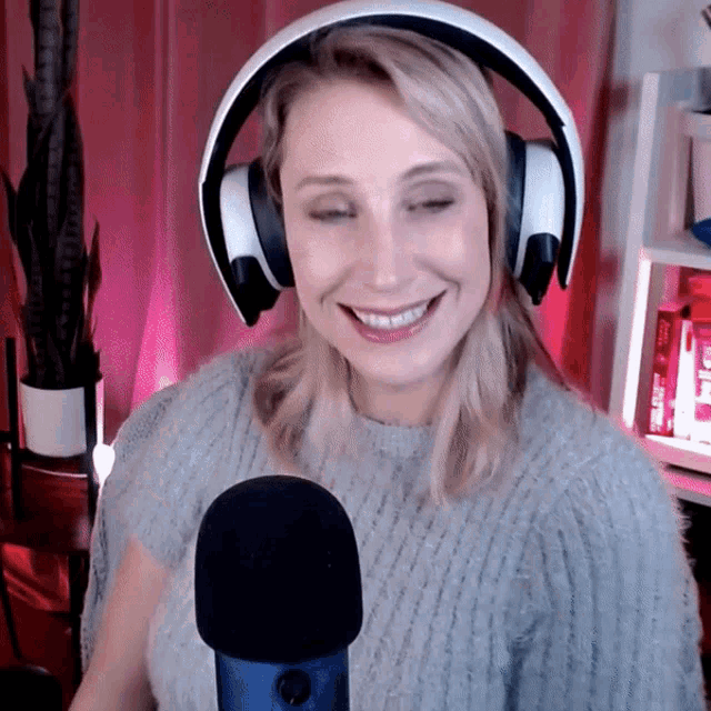 a woman wearing headphones and a grey sweater smiles in front of a microphone