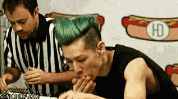 a man with green hair is eating a hot dog in front of a hd sign