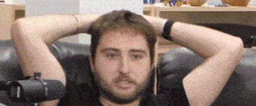 a man with a beard is sitting on a couch with his hands behind his head and a microphone in front of him .