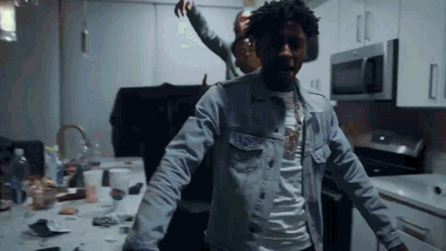 a man in a denim jacket is standing in a messy kitchen with his arms outstretched .