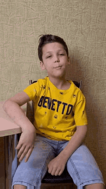 a young boy wearing a yellow benetton t-shirt is sitting in a chair
