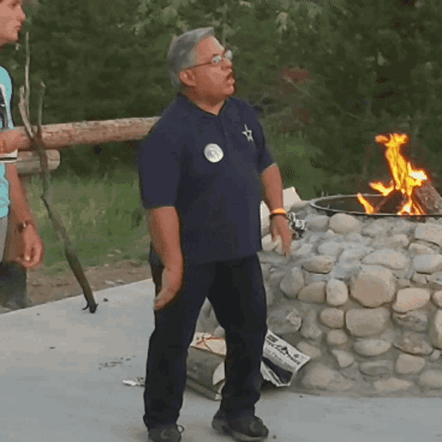 a man wearing a blue shirt with a star on the shoulder stands in front of a fire pit