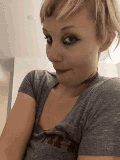 a woman wearing a choker and a grey t-shirt with the letter t on it is making a funny face
