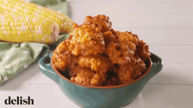 a blue bowl filled with corn fritters next to a corn on the cob .