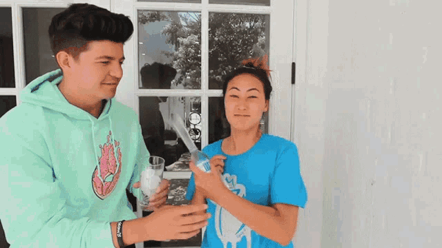 a man in a green hoodie is standing next to a woman in a blue shirt who is holding a glass
