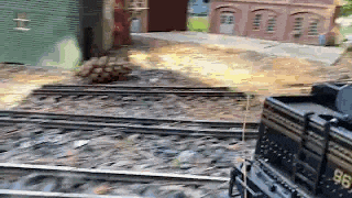 a train is going down the tracks with a building in the background and a brick building in the background .