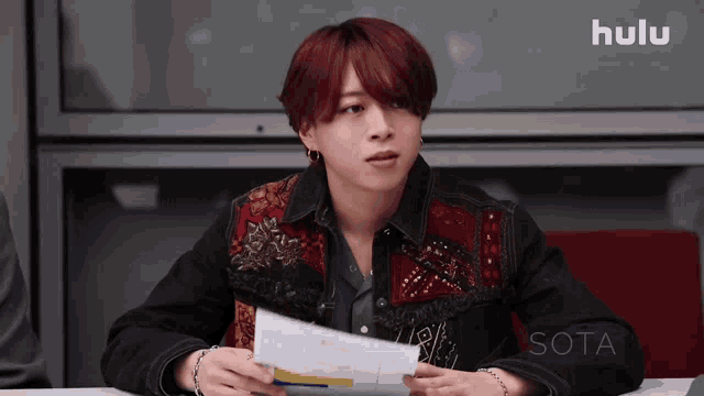 a man with red hair is sitting at a table holding a piece of paper in front of a screen that says hulu sota