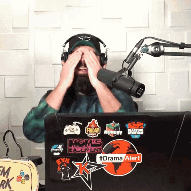 a man covering his eyes in front of a microphone with a laptop that says drama alert on it