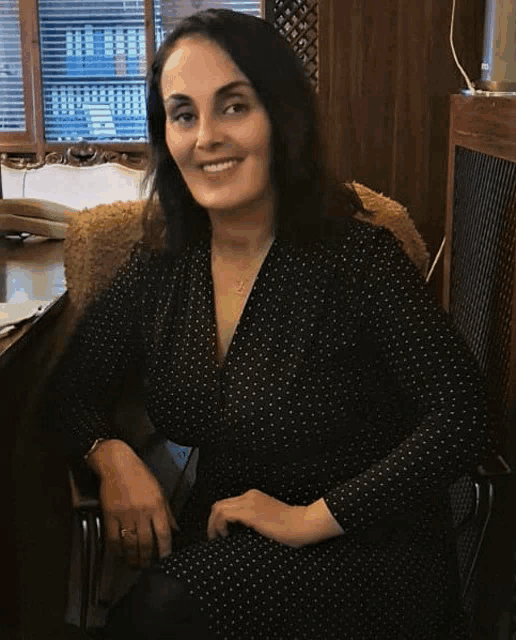 a woman in a black polka dot dress sits in a chair smiling