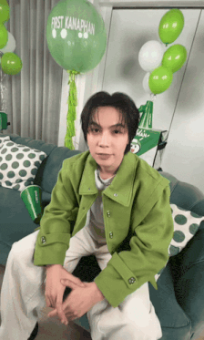 a man in a green jacket is sitting on a couch in front of a green balloon that says first kanaphan