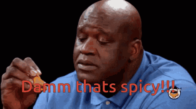 a bald man in a blue shirt is holding a piece of food and says " damn thats spicy " in red