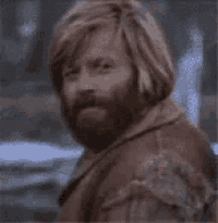 a man with long hair and a beard is wearing a brown jacket and looking at the camera .