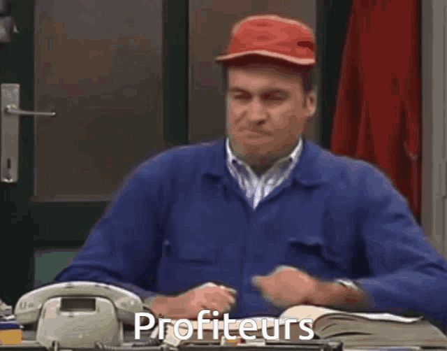 a man wearing a red hat is sitting at a desk with the word profiteurs written on his desk