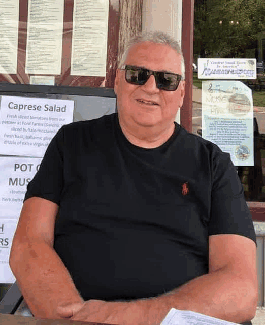 a man wearing sunglasses and a black shirt is sitting at a table in front of a sign for caprese salad .