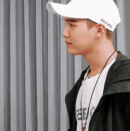 a young man wearing a white baseball cap and a black jacket is standing in front of a curtain .