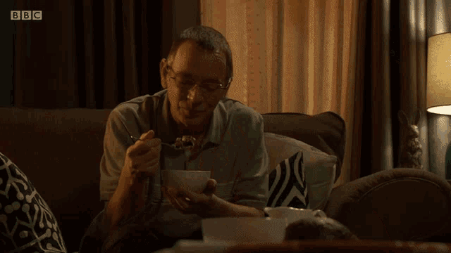 a man sits on a couch eating from a bowl with bbc written on the bottom left