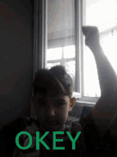 a boy is raising his fist in front of a window and the word okey is on the bottom of the picture