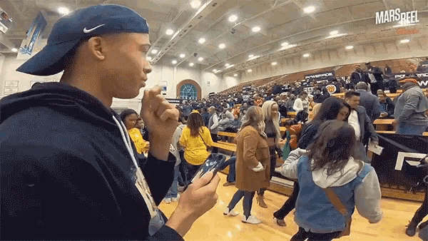 a man in a blue hat is looking at his phone in front of a crowd at a basketball game