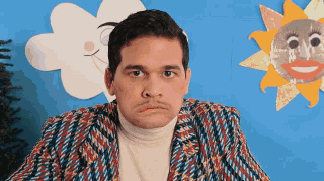 a man wearing a colorful jacket is sitting in front of a blue wall with a cloud and sun on it