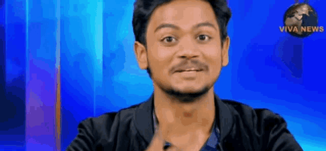 a man in a black jacket is making a funny face in front of a blue background that says viva news .